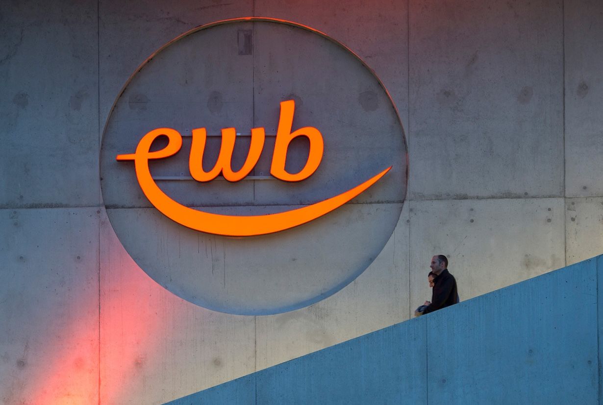 orange leuchtendes Logo der Energie Wasser Bern mit den Buschstaben ewb an einer Beton Wand und einer Treppe, bei der zwei Leute hinunterlaufen